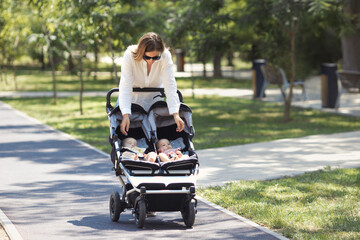 Lifestyle young casual caucasian mother with two little twin girls in a double baby stroller walks in city the park  - obrazy, fototapety, plakaty