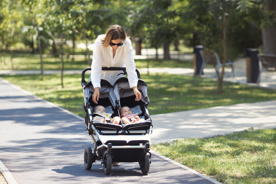 Lifestyle young casual caucasian mother with two little twin girls in a double baby stroller walks in city the park 