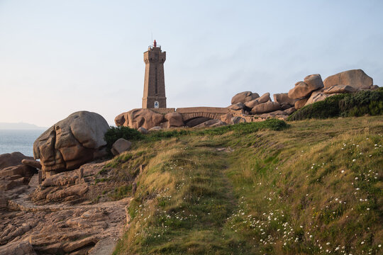 The lighthouse Men Ruz with bridge towering the extraordinarily colored and fantastically shaped rock formations at the walking trail dotted with wild flowers - pink granite coast in Brittany, France
