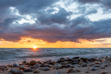 Sunset over ocean. Dark yellow sun set with sun rays touching the rough turquoise water along a rocky beach.  The sky is full of stormy dark clouds.