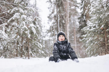 Fototapeta na wymiar Happy teenager boy sitting on snow in winter forest. Child having fun outdoors. Joyful adolescent playing in snow at snowfall. Laughing smiling kid walking in winter park in cold weather