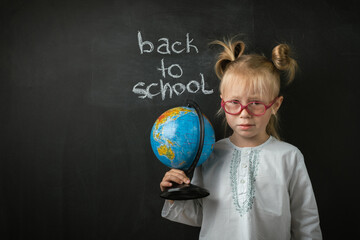 Fototapeta premium Portrait of little caucasian girl wearing glasses. Elementary school student holding the globe on the black school board background. Back to school concept