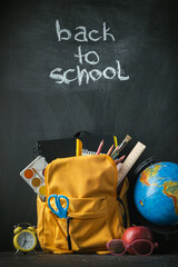 Yellow backpack with school supplies next to the globe, red apple and glasses on the black school board background. Back to school concept on September day