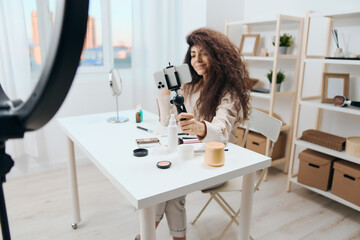 Enjoyed tanned handsome curly Latin beauty blogger in linen shirt record live with subscribers in home white interior. Copy space Mockup Banner. Influencer record video review blog using smartphone