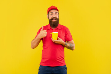 Naklejka premium Happy bearded man in casual red cap and tshirt giving thumb holding mug yellow background, coffee