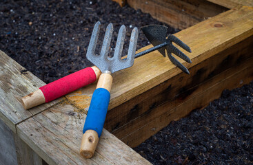 Weed fork and double sided hoe and rake garden hand tools on wooden vegetable planter box edge.