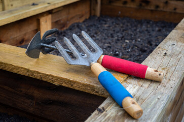 Weed fork and double sided hoe and rake garden hand tools on top of freshly planted wooden vegetable planter box. - obrazy, fototapety, plakaty