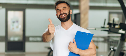 Portrait of personal trainer with clipboard showing thumb up at gym