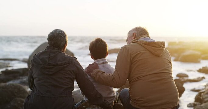 Grandfather and grandmother bonding with their little grandchild at sunset. A child and his grandparents at the sea sitting on rocks looking at the relaxing beach water outdoors together as a family