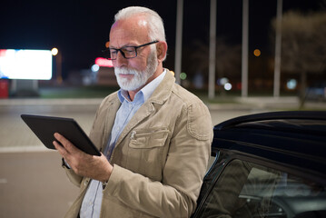 Fototapeta na wymiar Senior man standing outside of the car and using navigation system on parking lot at night 