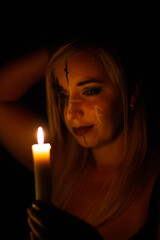 A beautiful blonde witch looks mysteriously at the candle flame. A woman with symbols on her face holds a candle in her hand in the dark. Halloween concept