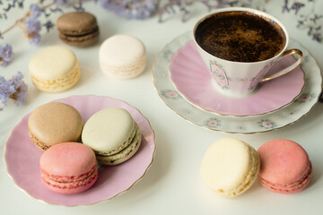 Mix of appetizing macarons and delicious coffee in porcelain cup on white table decorated with blurred flowers. Dry flowers. Greeting card.