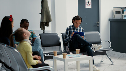 Asian patient filling in medical report at hospital reception lobby, sitting in waiting area to attend consultation appointment. Diverse patients waiting in healthcare facility.