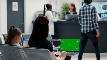 Mother and little girl using laptop with greenscreen in waiting room, holding computer at hospital reception. Woman looking at chroma key template with isolated mockup display and blank copyspace.