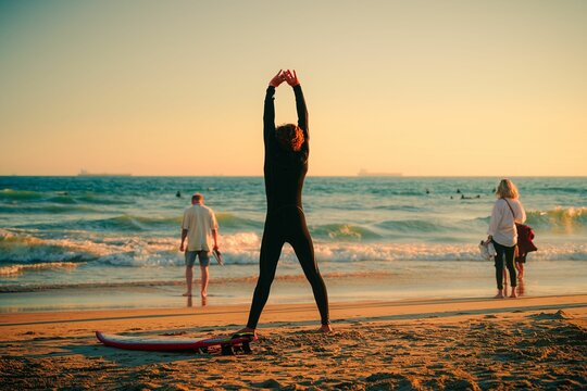 Slim surfer stretching at the beach with foamy waves at sunset