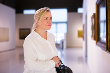 Portrait of attentive senior woman wearing in white, visiting painting exhibition