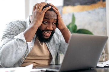 Irritated angry stressed african american man, sit at desk in the office, using laptop for online...