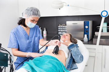 Confident female doctor performing radiofrequency lifting procedure on abdomen of mature woman