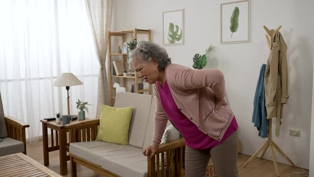 asian senior lady is feeling pain for she sprained her lower back while she is walking into the living room at home and supporting herself with sofa armrest