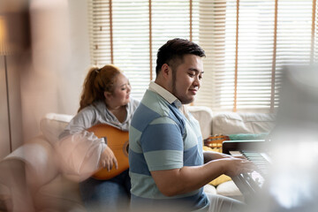 Fototapeta premium Young Asian chubby couple singing and playing acoustic guitar and piano together. Man and woman enjoying musical instrument. People in a band practicing in the house