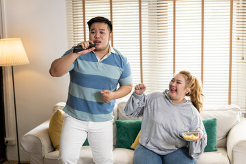 Young Asian chubby couple singing and watching tv on the couch. Man and woman enjoying a fun time together at home. People laughing and smiling together