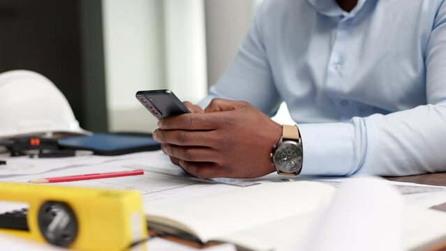 Closeup of the hands of an engineer or architect on a phone typing, reading email or on social media. Focused African guy using apps on a mobile, web surfing online, and searching for information.