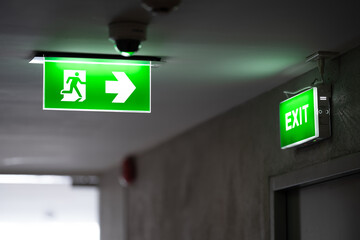 A green fire exit sign is placed on the ceiling along the dimly lit corridor and there is green...