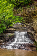 Obraz premium A small, beautiful waterfall splashes over a cliff and into a rocky forest canyon at McCormick’s Creek State Park, the first state park in Owen County, Indiana.