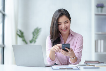 Beautiful Asian businesswoman is using her smartphone to chat with customers in a relaxed workplace with a smile and a good mood.