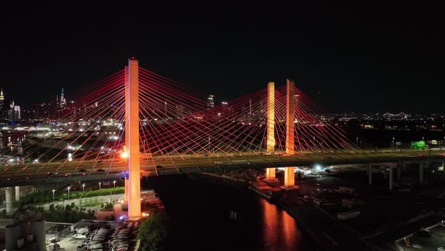 An aerial view of the illuminated cables and concrete towers of the Kosciuszko Bridge, lit up by light emitting diodes. The NYC skyline is in the background as the camera dolly out and boom up.
