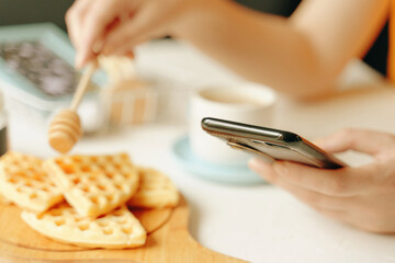 Morning romantic pastime. Online communication over lunch. Woman writes messages on cell phone and breakfast. Belgian waffles on desk, cup of coffee and honey stick on white table.