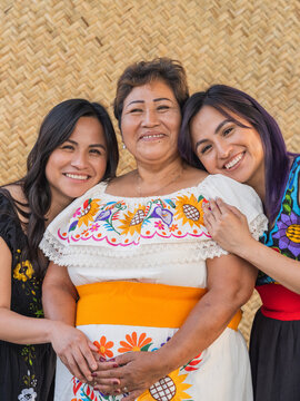 Vertical image of three Indigenous women smiling directly into the camera with a petate as the background 