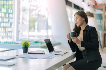 Obraz premium Smiling beautiful Asian businesswoman holding coffee cup and laptop working on documents graphs analyze charts using calculators at financial accounting concept office