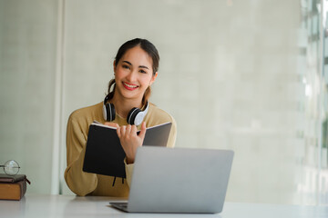 A young Asian student studying online using headphones and a laptop takes notes in a notebook sitting at her desk at home.