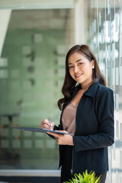 Attractive Asian businesswoman stands holding digital tablet next to glass flap in modern office.