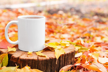 White ceramic mug close-up on a wooden background with colorful leaves. Autumn comfort, hot tea in a cup with copy space.