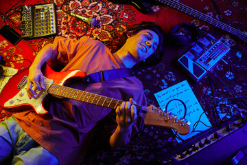 Top view shot of young musician playing guitar while lying on carpet lit by neon light