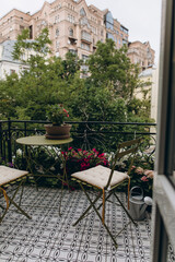 a coffee table and two chairs on an open balcony with a city view. blooming flowers in pots decorate the balcony