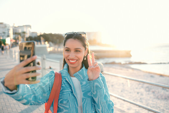 Smiling, trendy and fun woman, tourist or influencer taking selfie with peace sign gesture on phone for social media, video call or photo. Exploring, traveling and sightseeing at promenade outdoors
