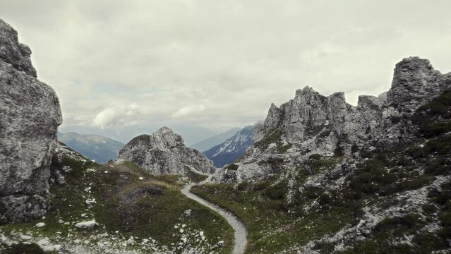 Cinematic aerial. Flying along a small trail with steep rock formations around and grassy ground near 11er peak. A beautiful mountain range scenery and a valley in the background. Stubai alps, Austria