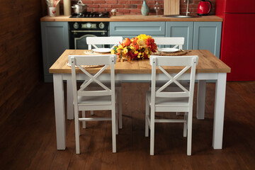 Stylish dining room with wooden table and chairs. Cozy cuisine decorated with fall decor and table setting flowers and pumpkins. Interior design scandinavian kitchen with utensils, dishes, plates.	