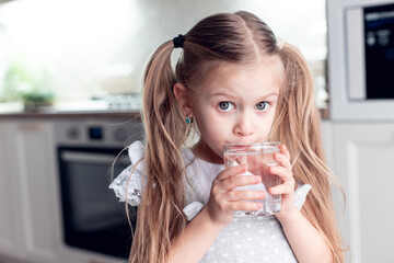 cute little girl drinks clean fresh water from a transparent glass at home in the kitchen. useful...