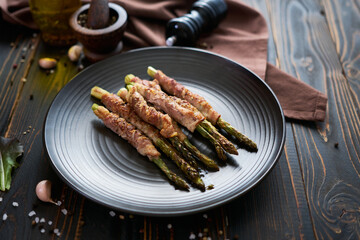 baked aspargus covered with bacon on a black ceramic plate on wooden kitchen table