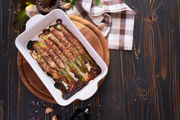 ceramic baking dish with baked aspargus covered with bacon on wooden kitchen table
