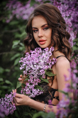 a beautiful girl with makeup and hair styling in underwear stands in a garden with lilacs on a summer evening