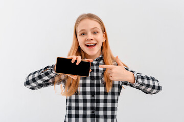 Portrait of smiling teen girl pointing finger at mobile phone screen, showing online application, standing over white background