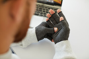 Young man who suffers from rheumatoid arthritis wears pair of warm fingerless grey textile compression gloves for relieving pain in his hands and wrists while working on notebook PC at his office desk - obrazy, fototapety, plakaty