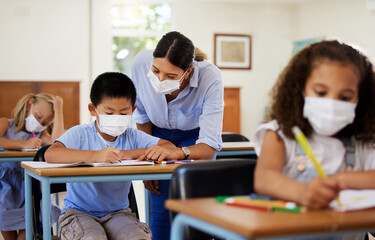 Fototapeta premium Covid, education and learning with a teacher wearing a mask and helping a male student in class during school. Young boy studying in a classroom with help from an educator while sitting at his desk