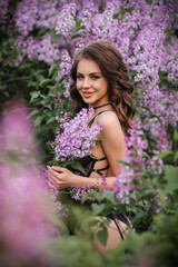 a beautiful girl with makeup and hair styling in underwear stands in a garden with lilacs on a summer evening