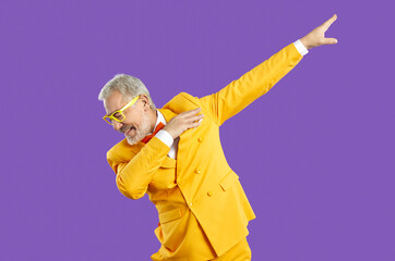 Funny happy confident handsome senior man in yellow suit and eyeglasses doing dab moves. Cheerful eccentric mature guy wearing trendy outfit dancing isolated on purple background. Fashion concept
